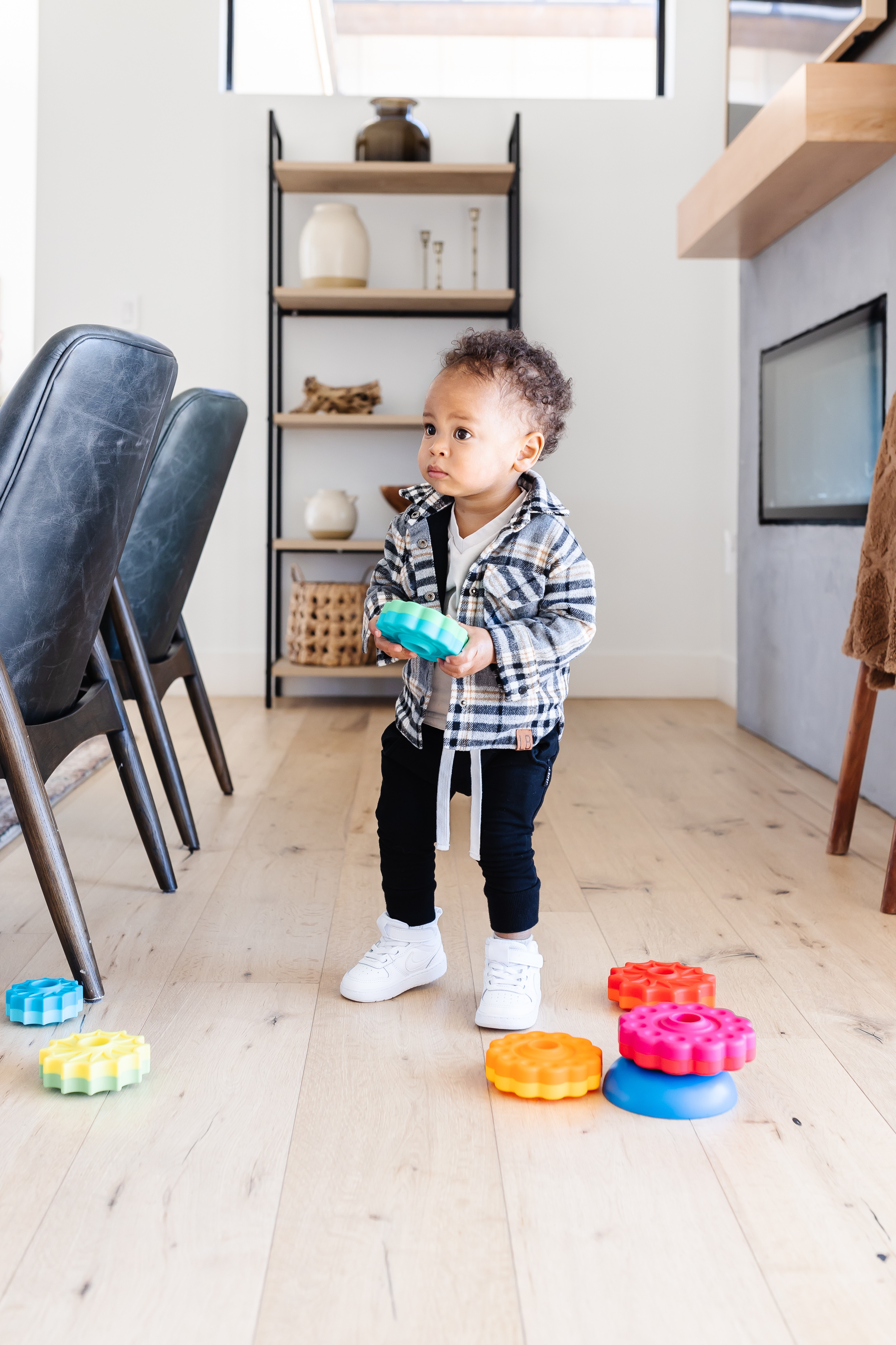 Child standing in a room holding a toy, with colorful toys on the floor, a shelf with decorative items, and chairs nearby.