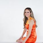 Smiling woman in orange dress sitting at a table with color swatches and magazines, against a plain white background.