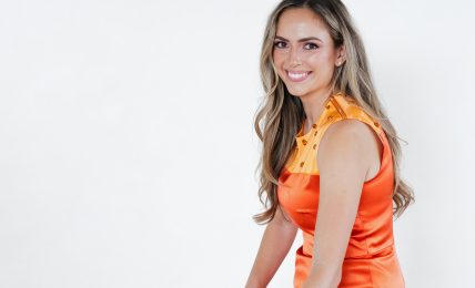 Smiling woman in orange dress sitting at a table with color swatches and magazines, against a plain white background.