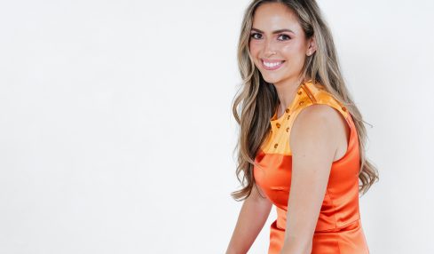 Smiling woman in orange dress sitting at a table with color swatches and magazines, against a plain white background.