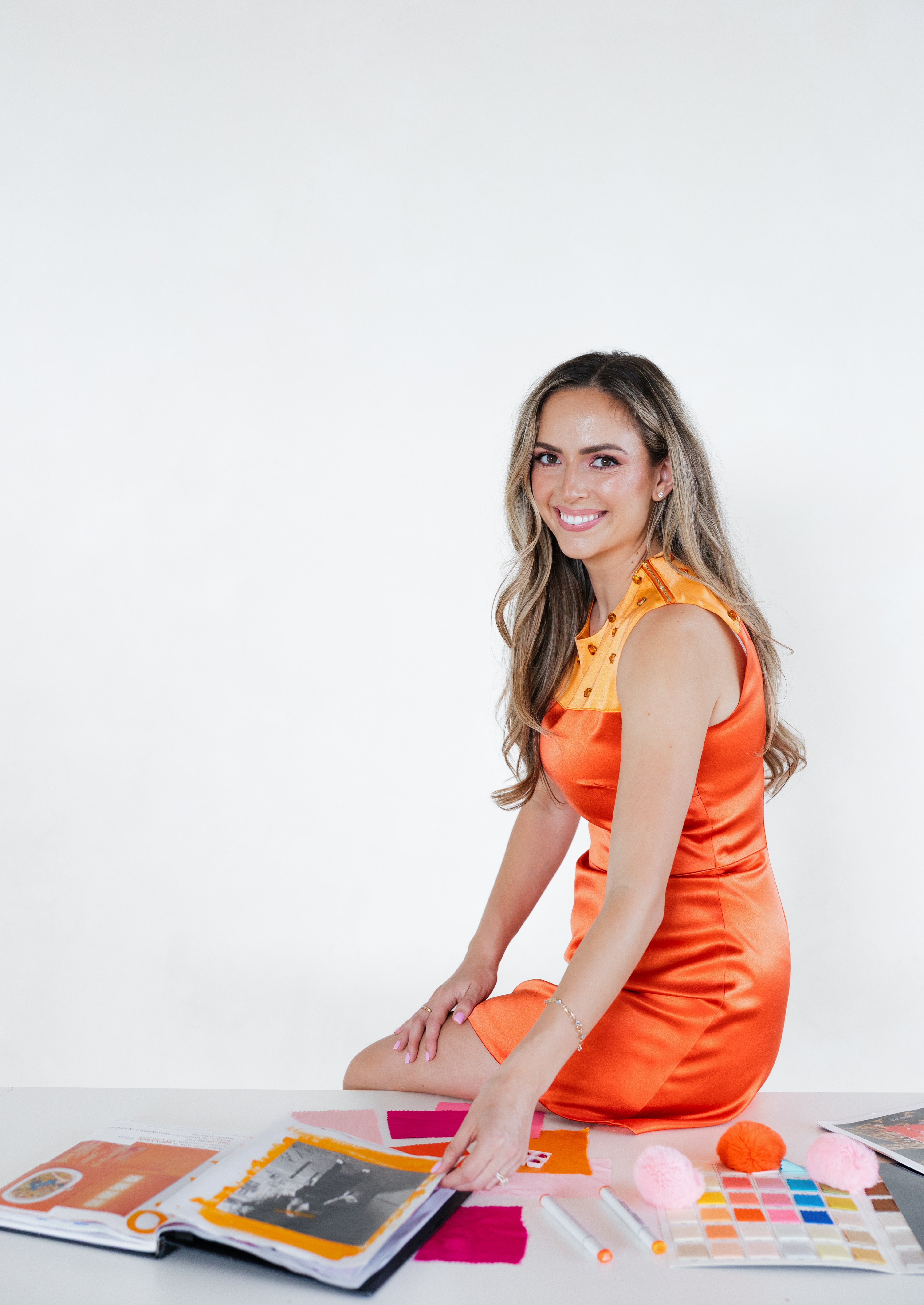 Smiling woman in orange dress sitting at a table with color swatches and magazines, against a plain white background.