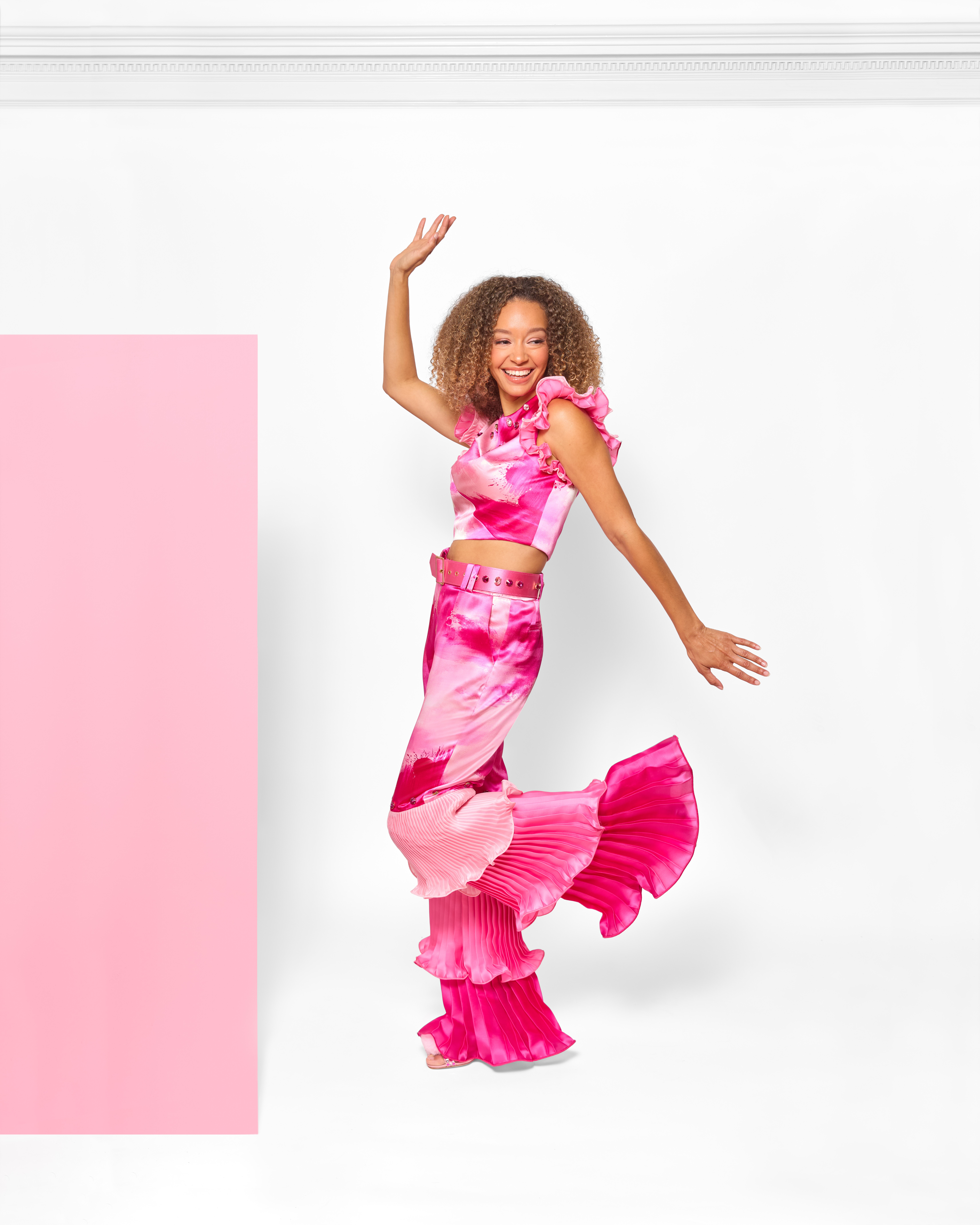 Young girl dancing with curly hair, wearing a pink tie-dye outfit, smiling, against a white background with a pink rectangle.