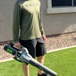 Person standing outdoors on grass holding a leaf blower, wearing a cap, long-sleeve shirt, and shorts.