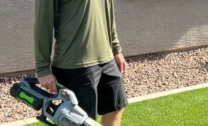 Person standing outdoors on grass holding a leaf blower, wearing a cap, long-sleeve shirt, and shorts.