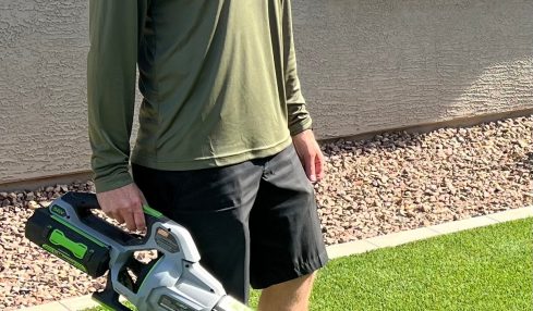 Person standing outdoors on grass holding a leaf blower, wearing a cap, long-sleeve shirt, and shorts.