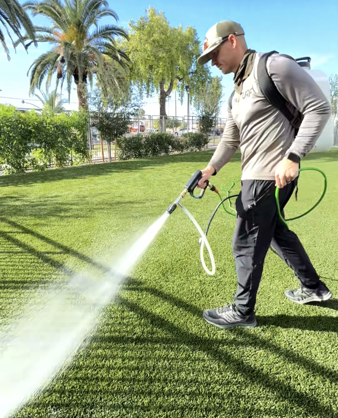 Person wearing a cap and backpack using a spray tool on grass in a park with trees and blue sky