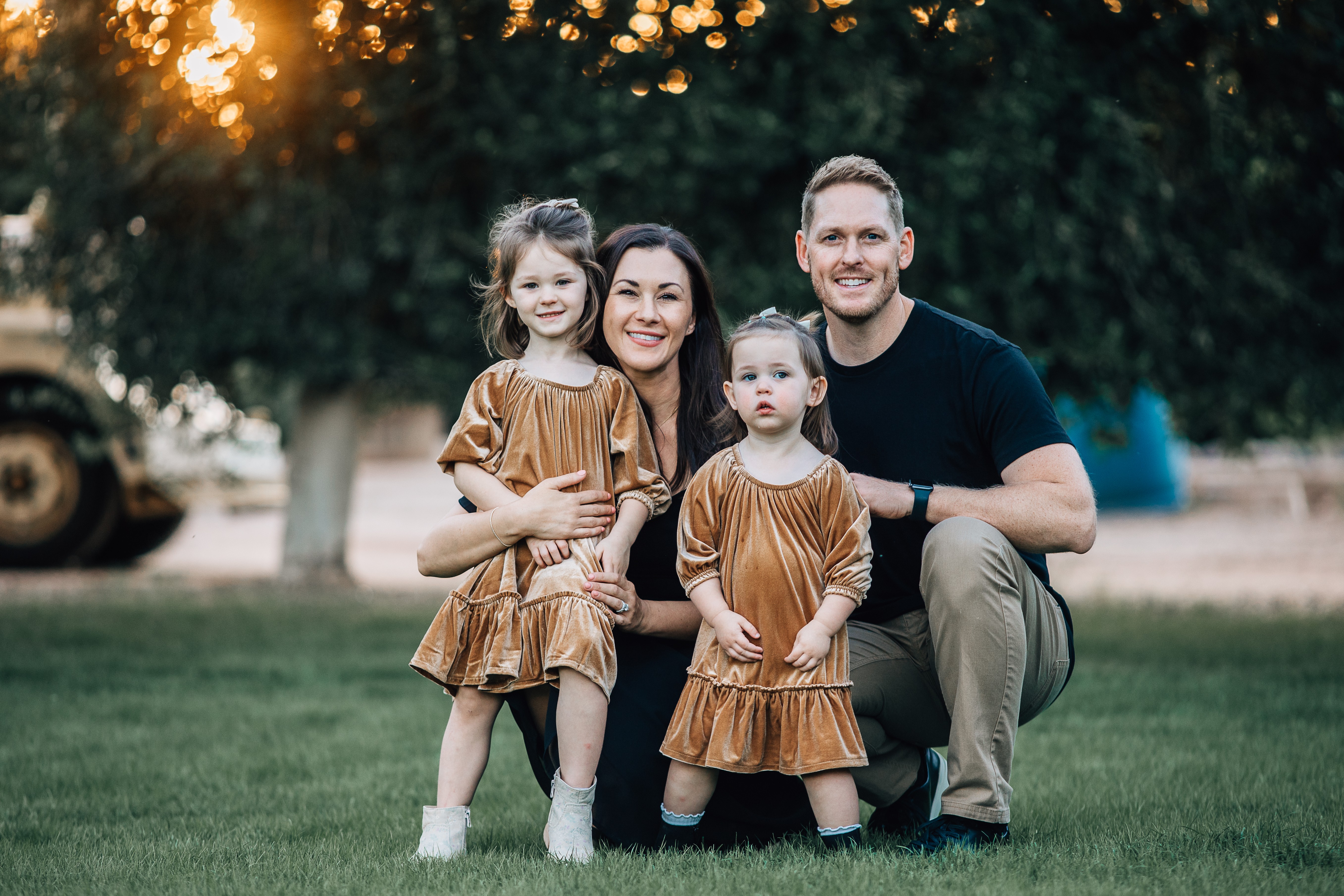 Family of four outdoors, two young girls and two adults, standing on grass with trees in background, sunset lighting.