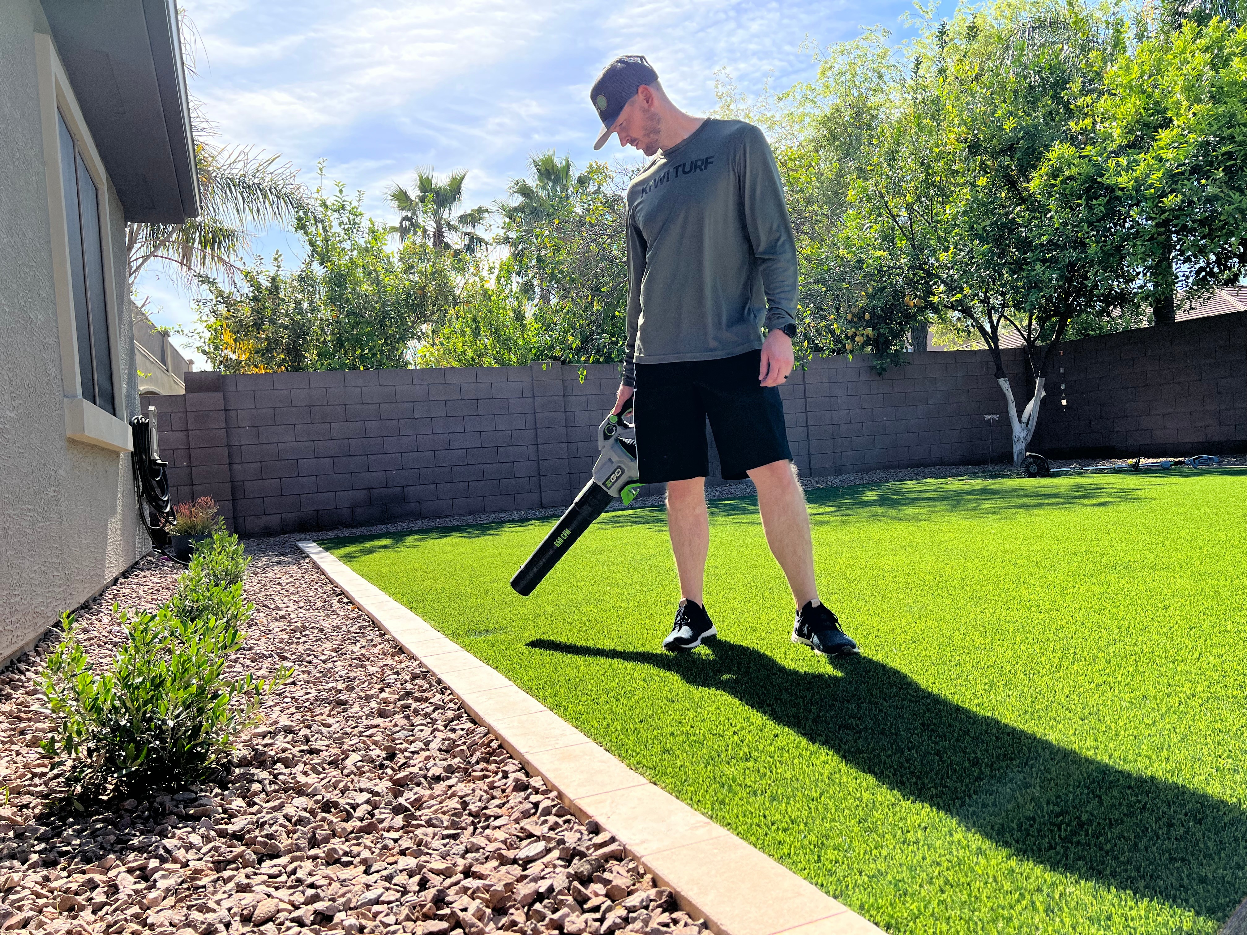 Person in gray shirt and black shorts using leaf blower on lawn, with trees and a brick wall in background.