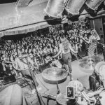 Band performs on stage with audience watching, musical instruments visible, in black and white, lively concert scene.