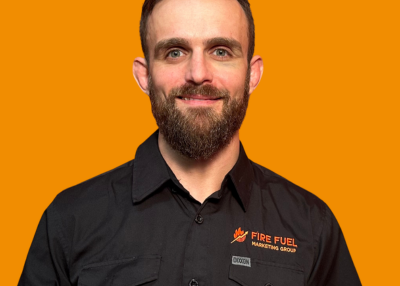 Portrait of a man with a beard and short hair, wearing a black shirt with a logo, against an orange background.