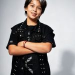 Young person with short dark hair smiling, arms crossed, wearing a black jacket and pants, standing against a light background.