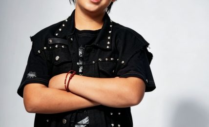 Young person with short dark hair smiling, arms crossed, wearing a black jacket and pants, standing against a light background.