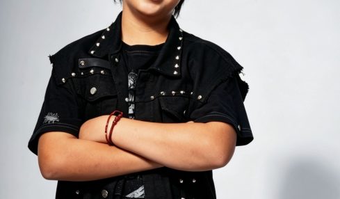 Young person with short dark hair smiling, arms crossed, wearing a black jacket and pants, standing against a light background.