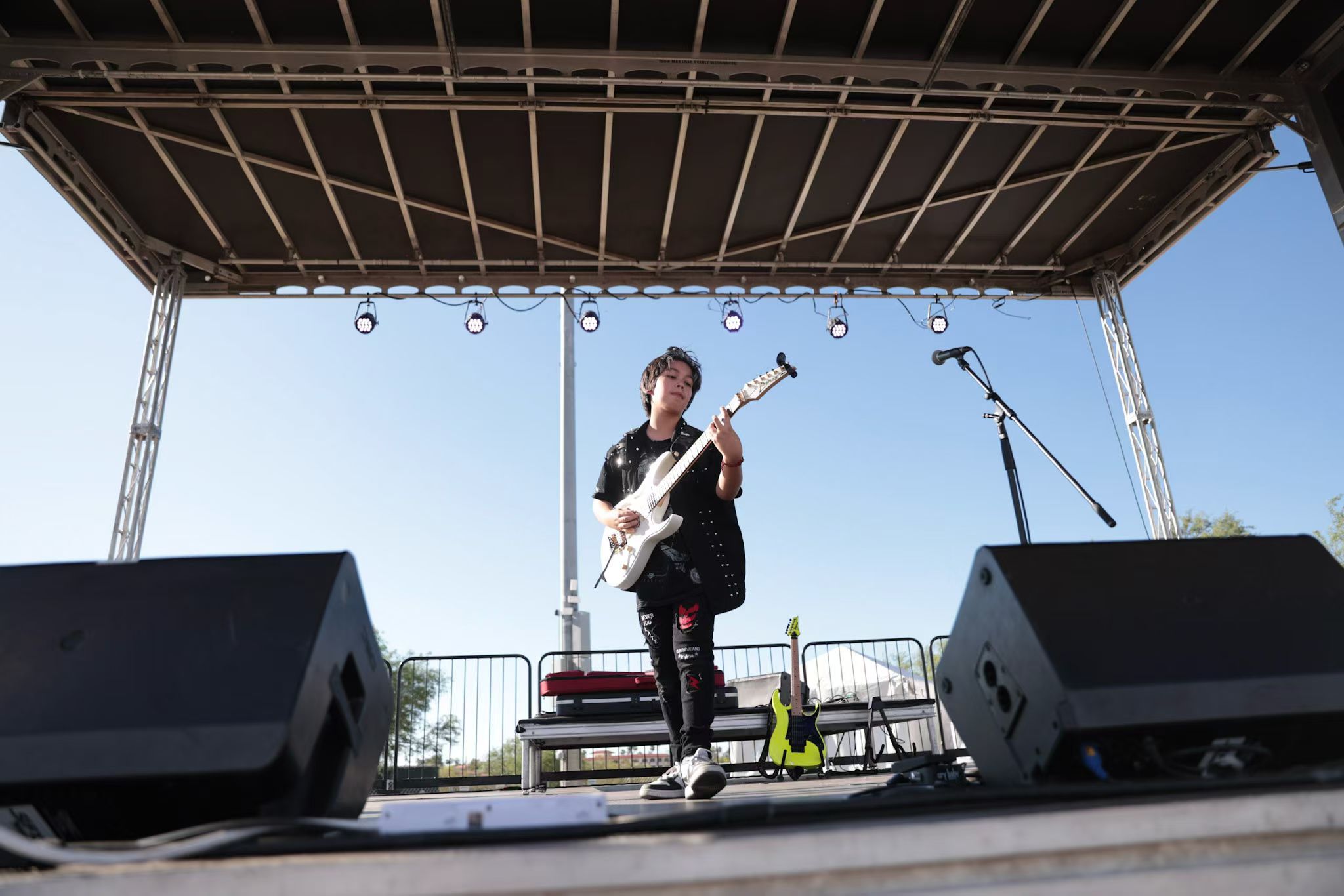 Person playing guitar on an outdoor stage with microphones and speakers, under a metal canopy, against a clear sky.