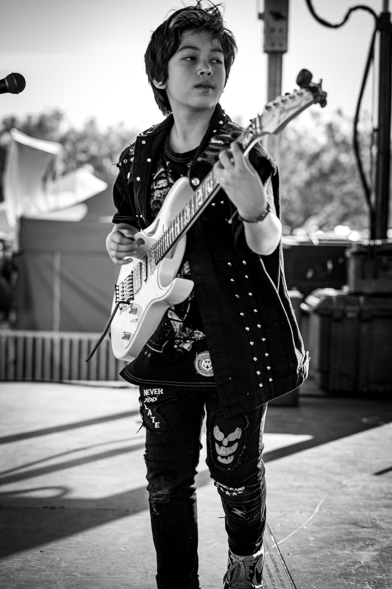 Young person playing electric guitar outdoors, focused on performance, with trees and equipment in background.