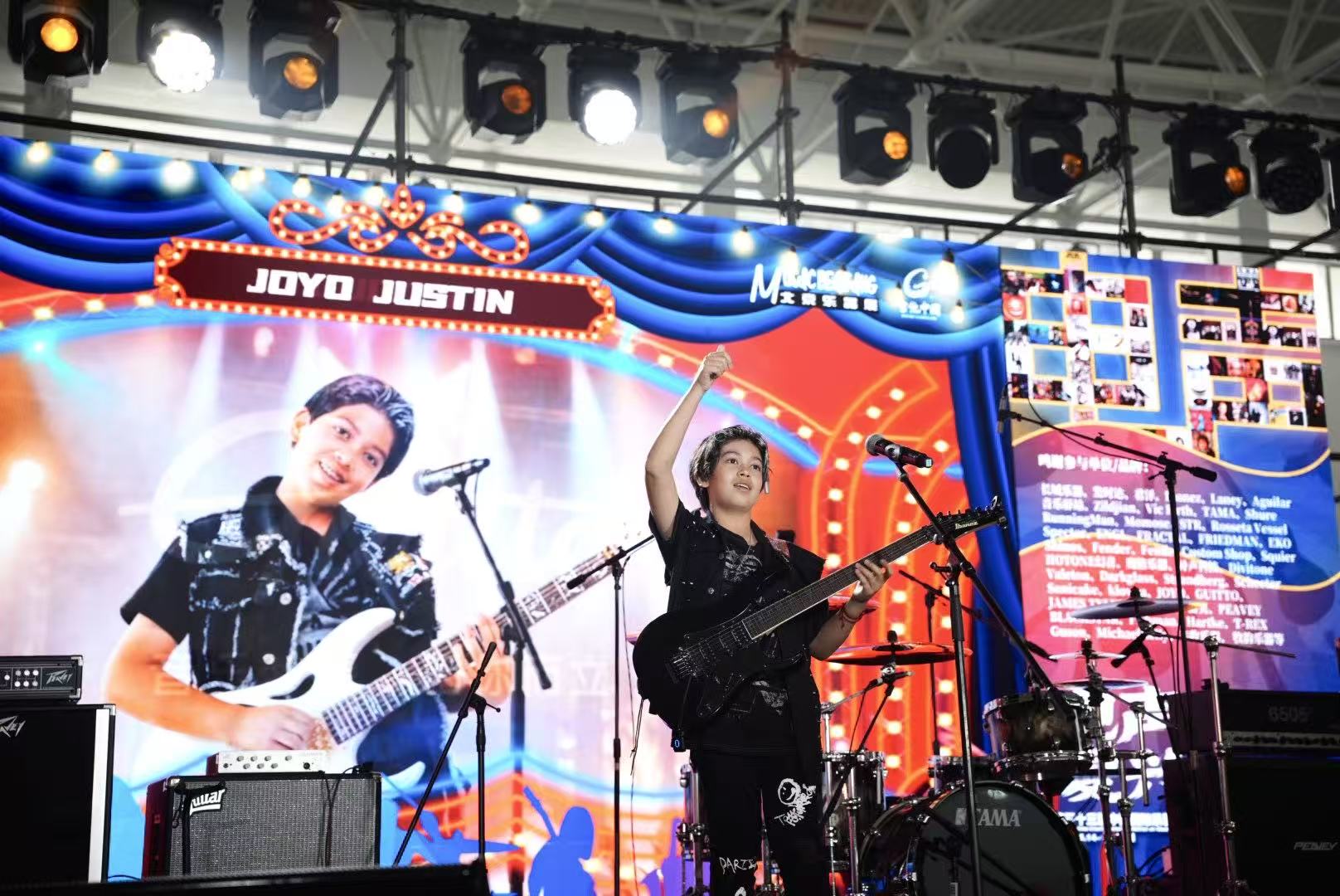 Two musicians perform on stage with colorful lights and a large screen behind them, one playing guitar and singing, the other singing.