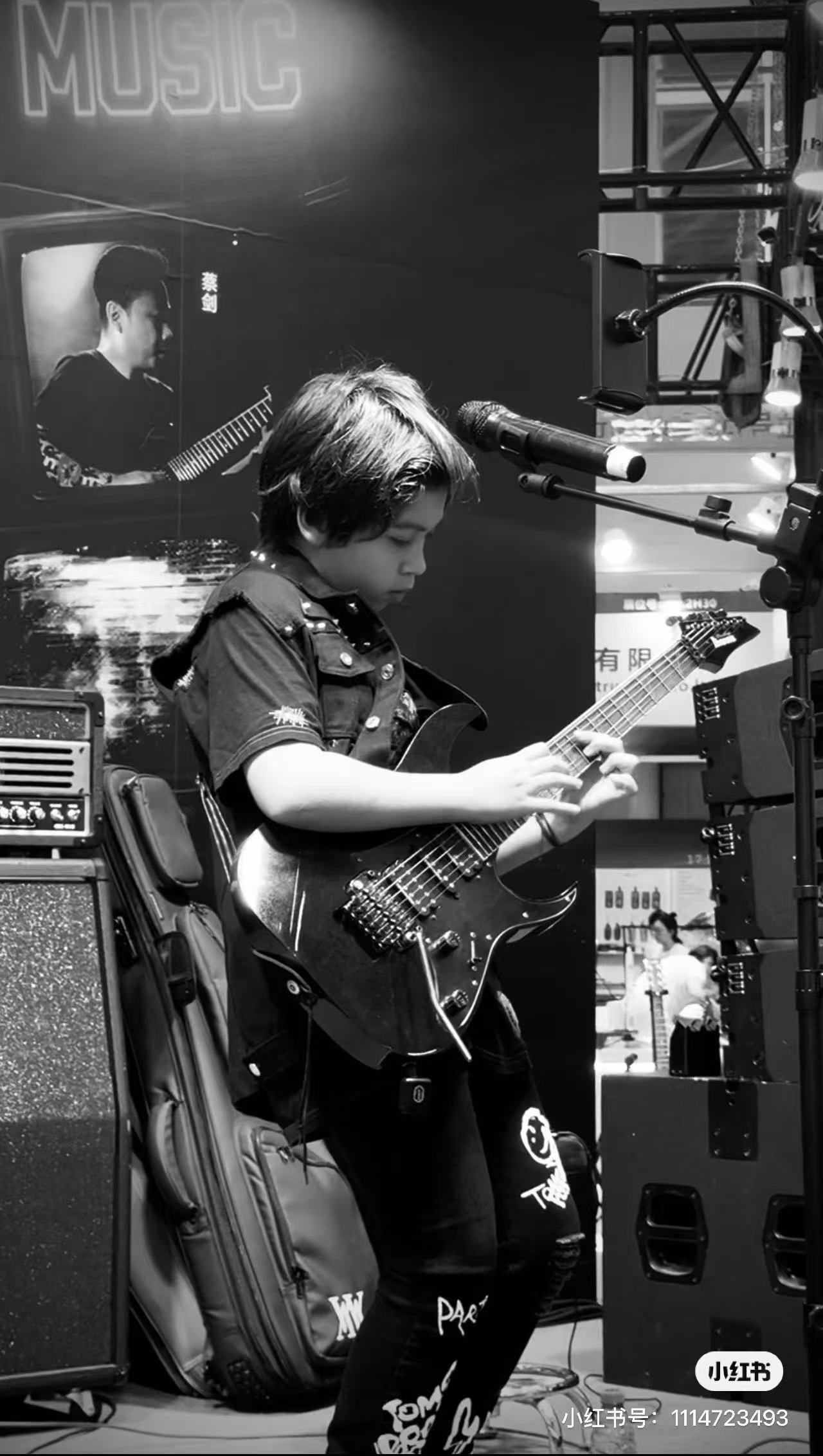 Young person playing an electric guitar on stage with a microphone, music equipment, and a large screen in the background.