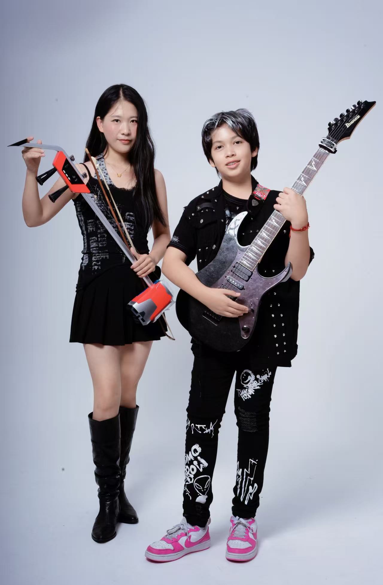 Two young girls holding toy musical instruments, one with a toy guitar and the other with a toy electric guitar, standing against a plain background.