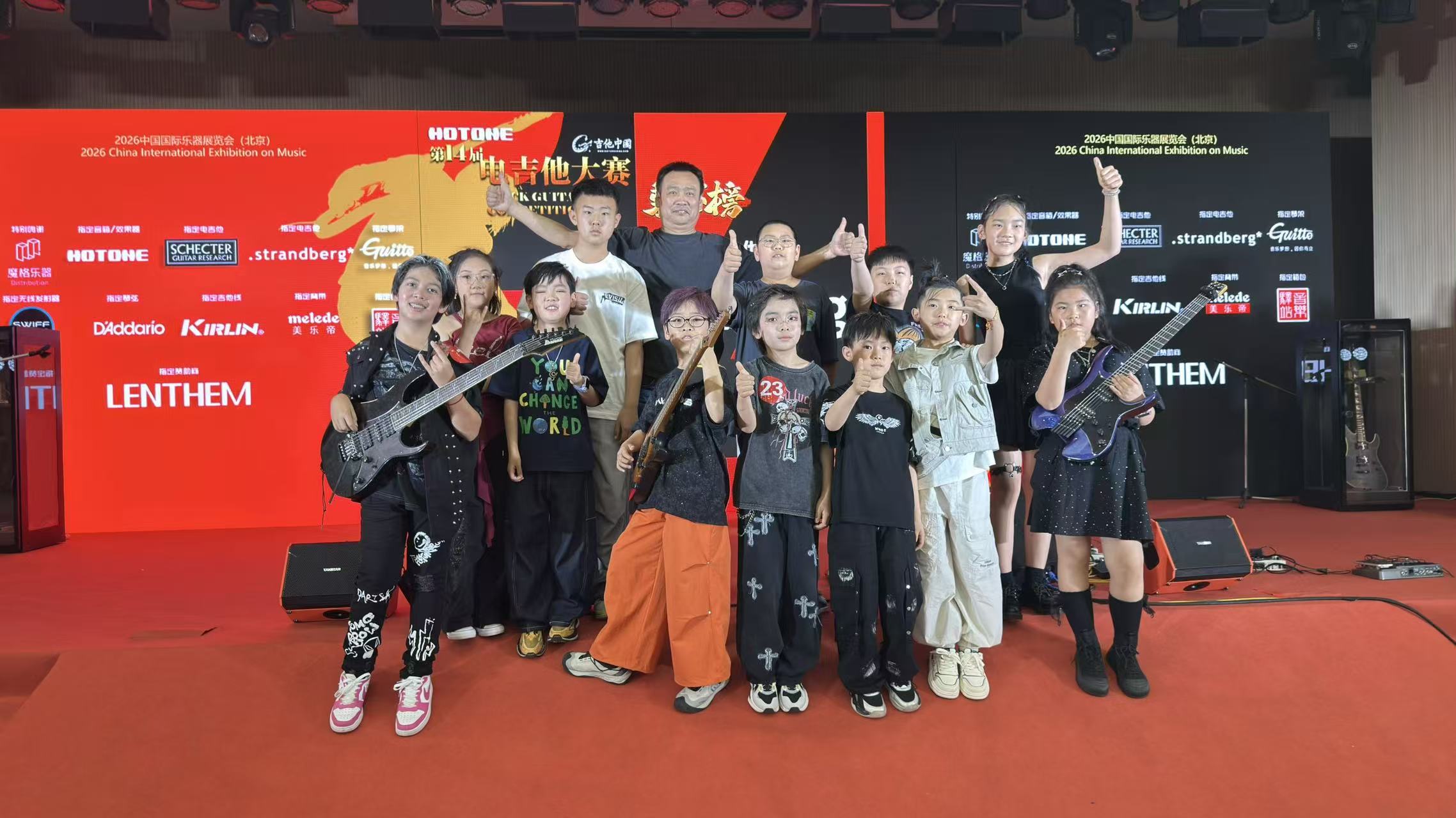 Group of children and adults on stage holding guitars, with red and black background banners, some raising hands.