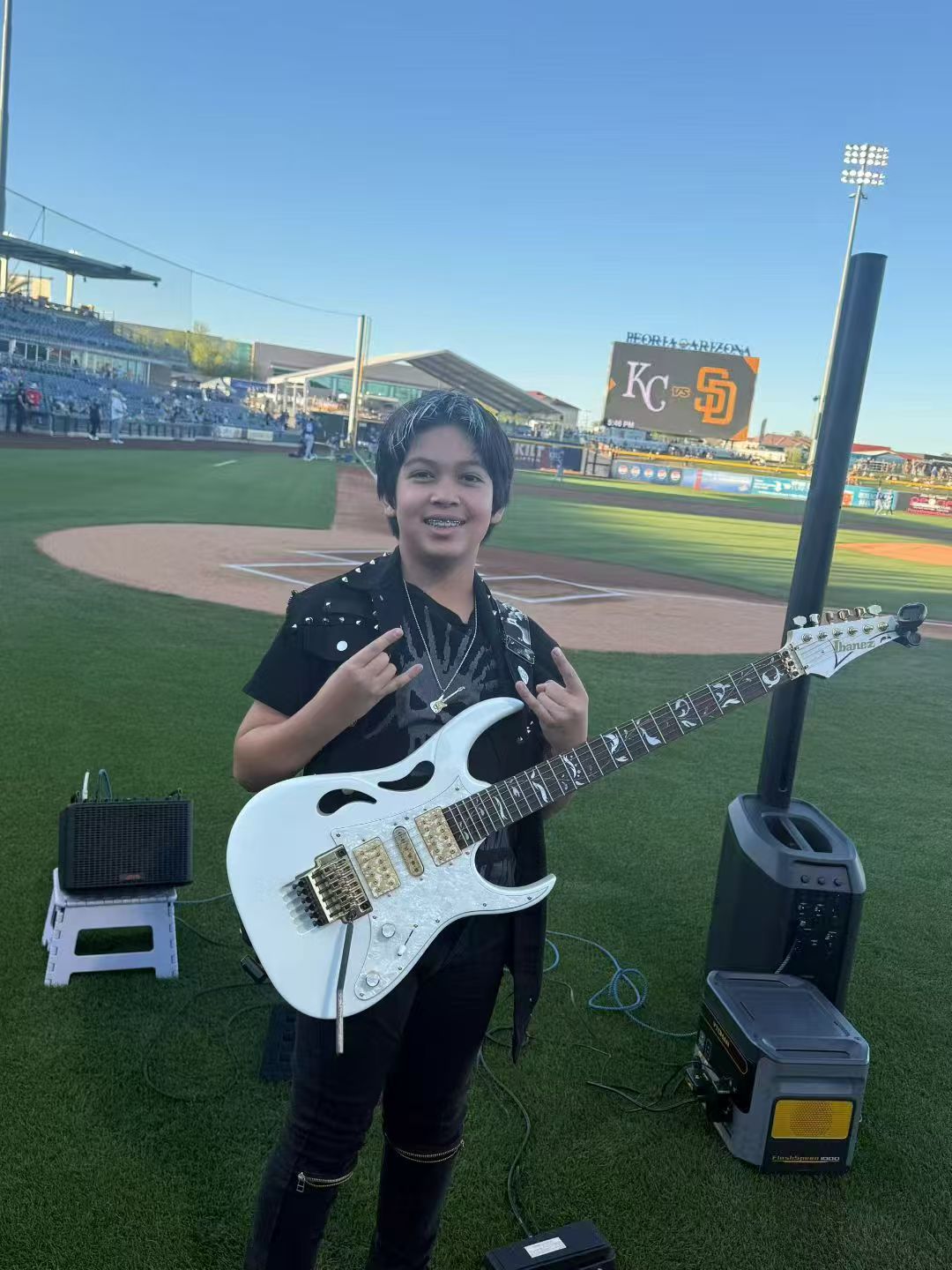 Young boy holding a white electric guitar on a baseball field, with stadium and scoreboard in background.