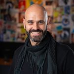 Bald man with beard smiling, wearing a black shirt and scarf, in front of a colorful collage background.