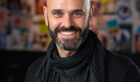 Bald man with beard smiling, wearing a black shirt and scarf, in front of a colorful collage background.