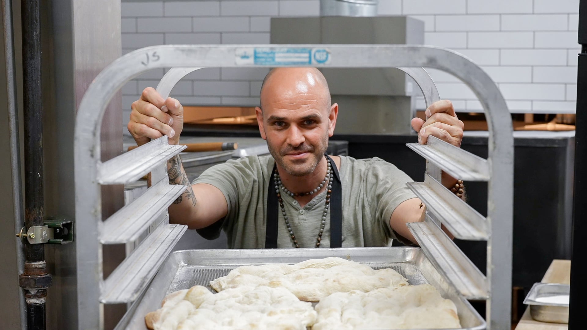 Man with a bald head and beard looking through a metal rack in a bakery or kitchen, with dough in front.