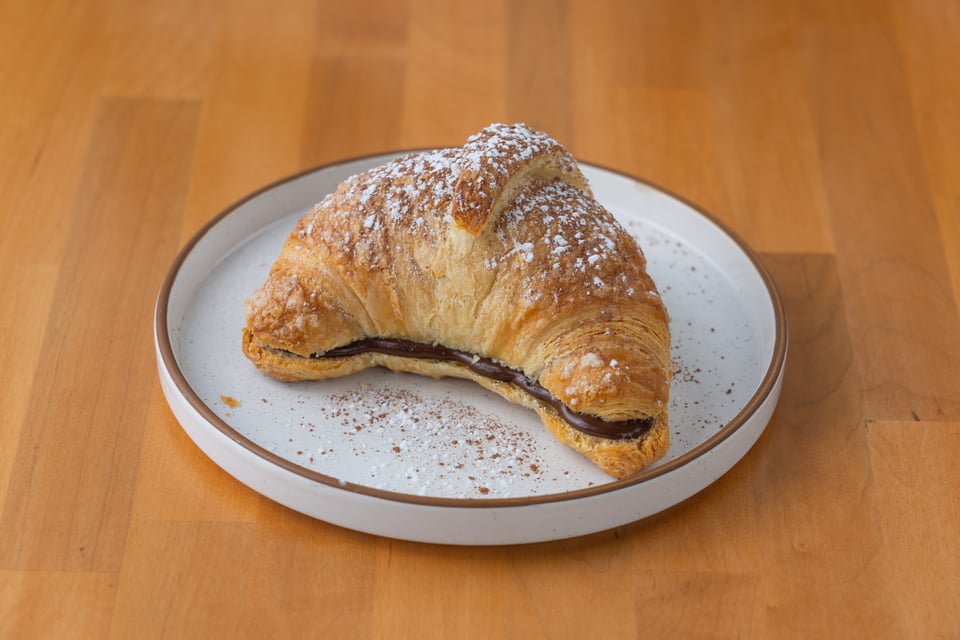 Croissant with chocolate filling on a white plate, dusted with powdered sugar, on a wooden surface.