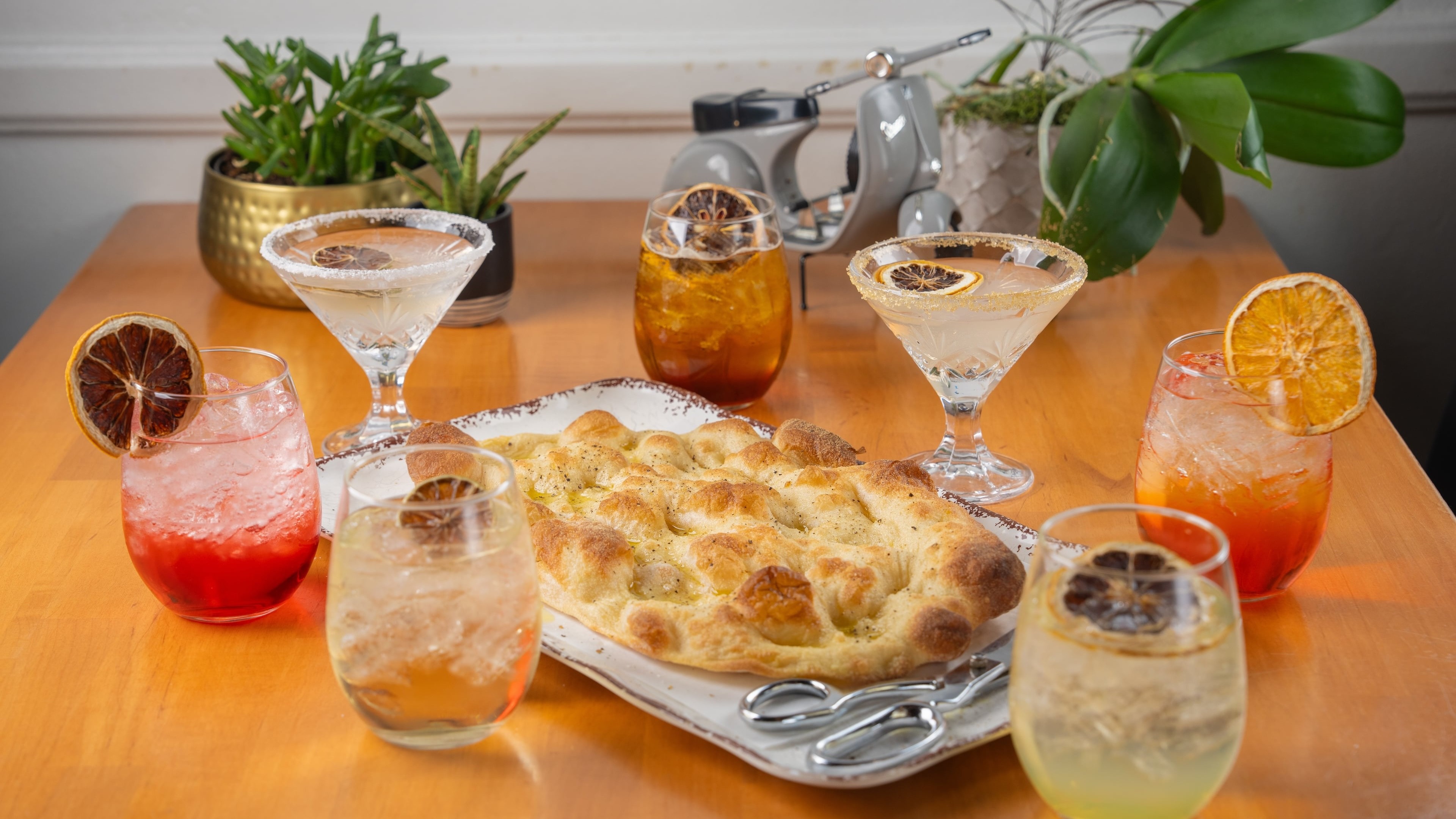 Table with a pizza, six drinks, and a potted plant in the background.