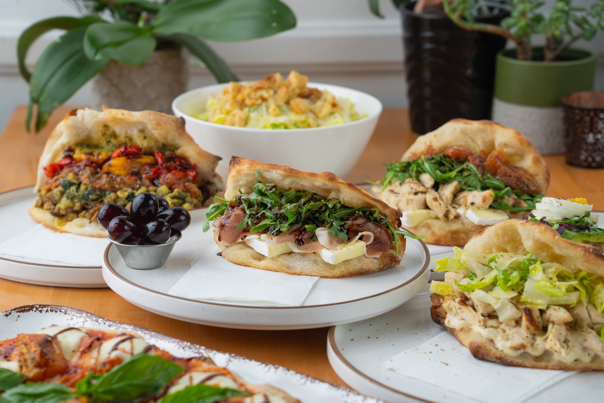 Sandwiches with various fillings, a bowl of rice, and plants in the background on a wooden table.