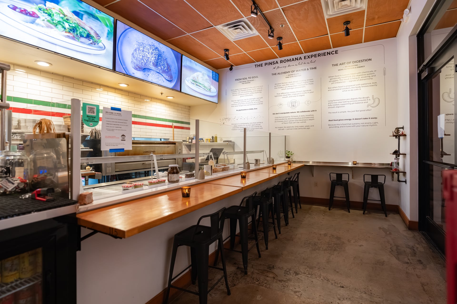 Empty counter with stools, menu on wall, and screens above in a restaurant or cafe setting.