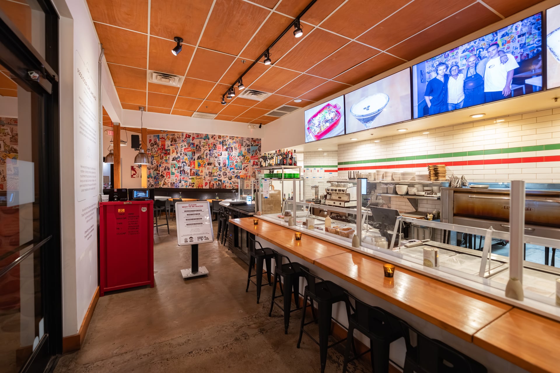 Empty restaurant with a long wooden counter, colorful wall art, and digital screens displaying images, with seating and food service area.