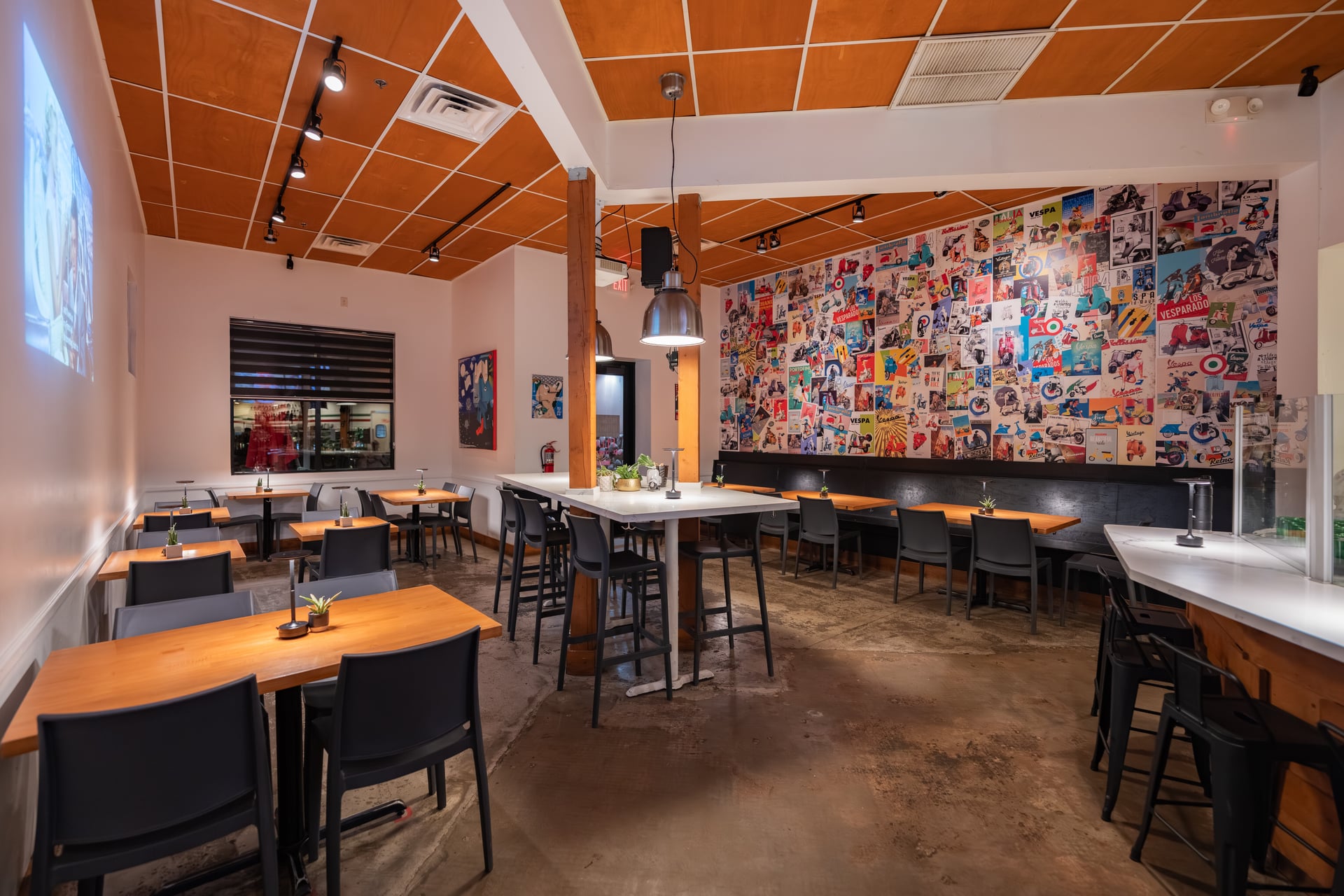 Interior of a restaurant with wooden ceiling, colorful collage wall, and black chairs around tables, illuminated by warm lighting.