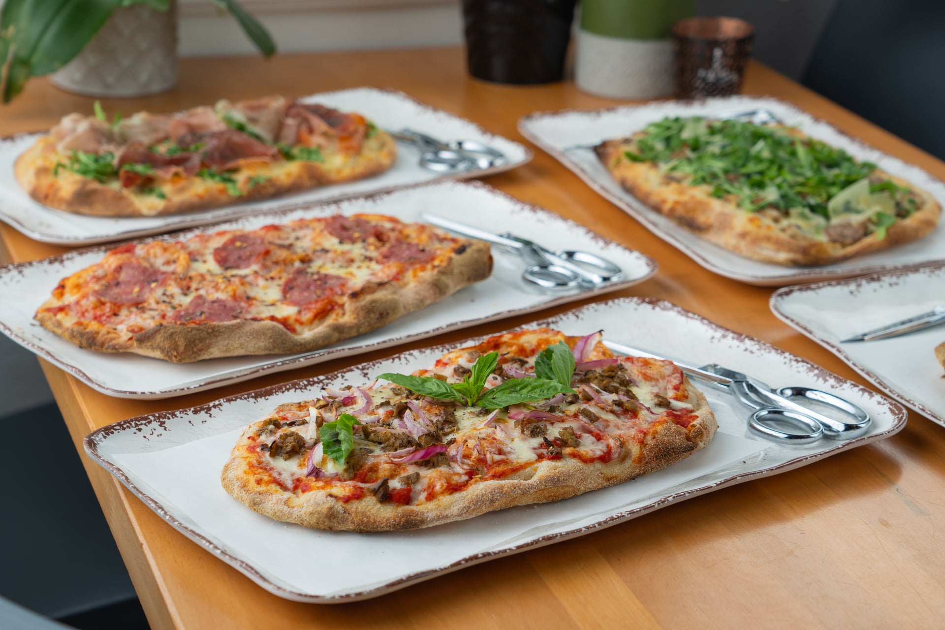 Three pizzas on white plates with silver utensils on a wooden table, toppings include vegetables and meats.