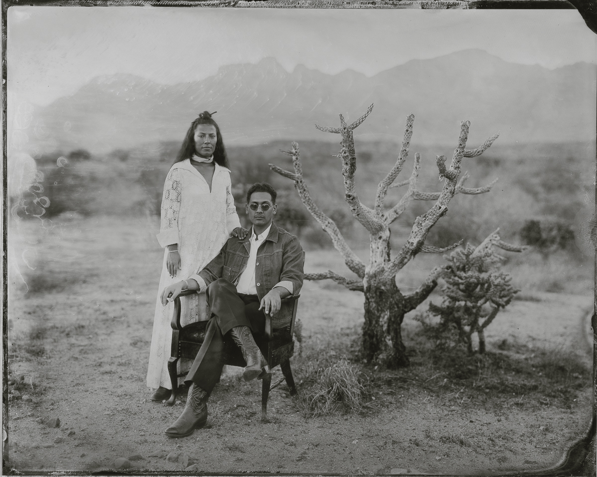 A woman and a man pose outdoors with a leafless tree, woman standing and man sitting on a chair, black and white photo.