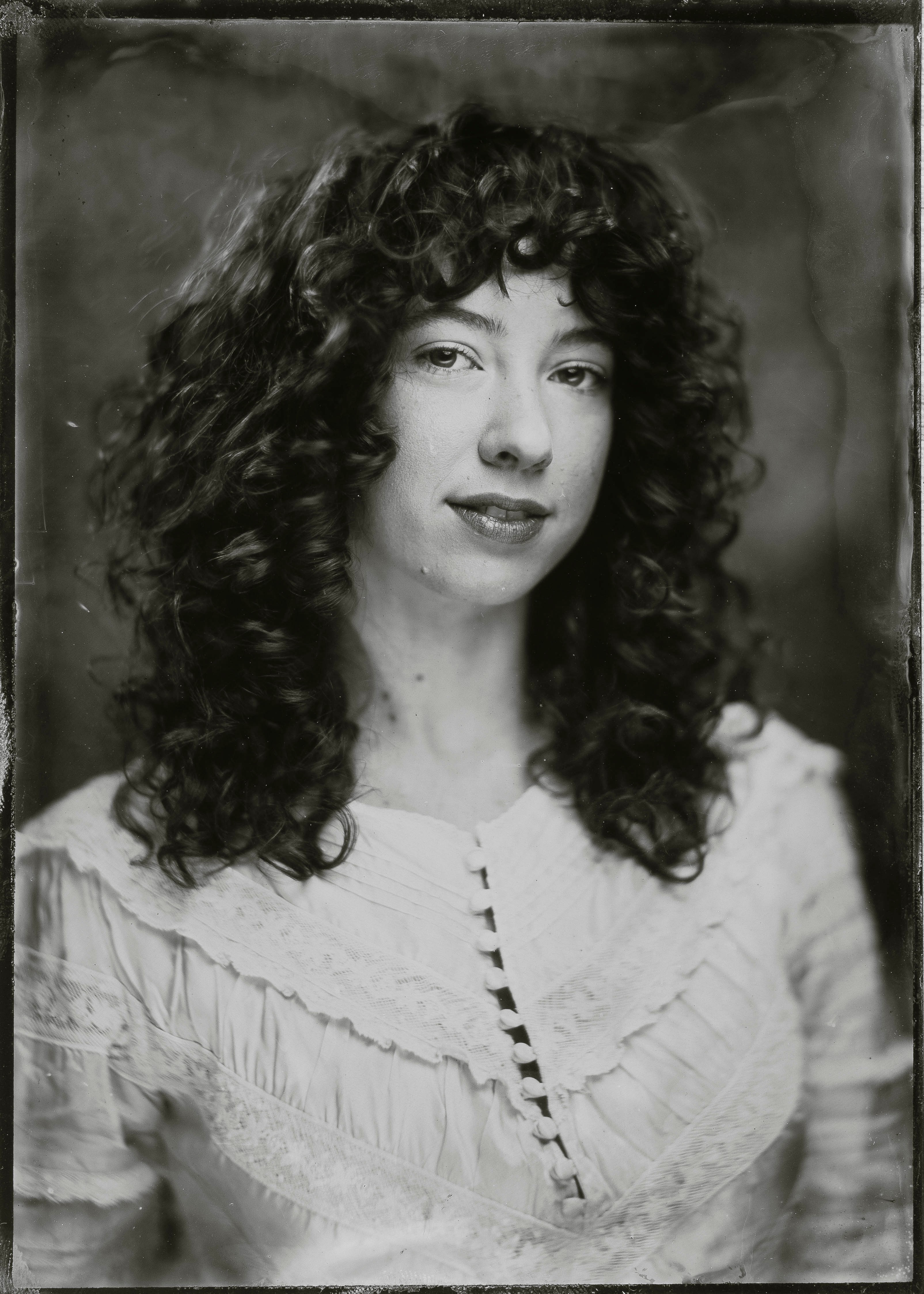 Black and white portrait of a woman with curly hair wearing a ruffled blouse, looking at the camera.