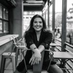 Woman sitting on a chair in a cafe, smiling, with large windows and outdoor seating visible behind her.
