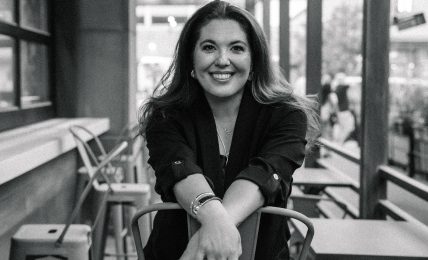 Woman sitting on a chair in a cafe, smiling, with large windows and outdoor seating visible behind her.