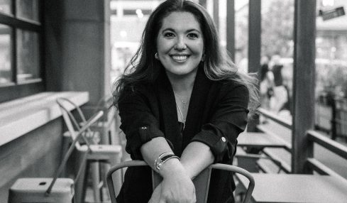 Woman sitting on a chair in a cafe, smiling, with large windows and outdoor seating visible behind her.