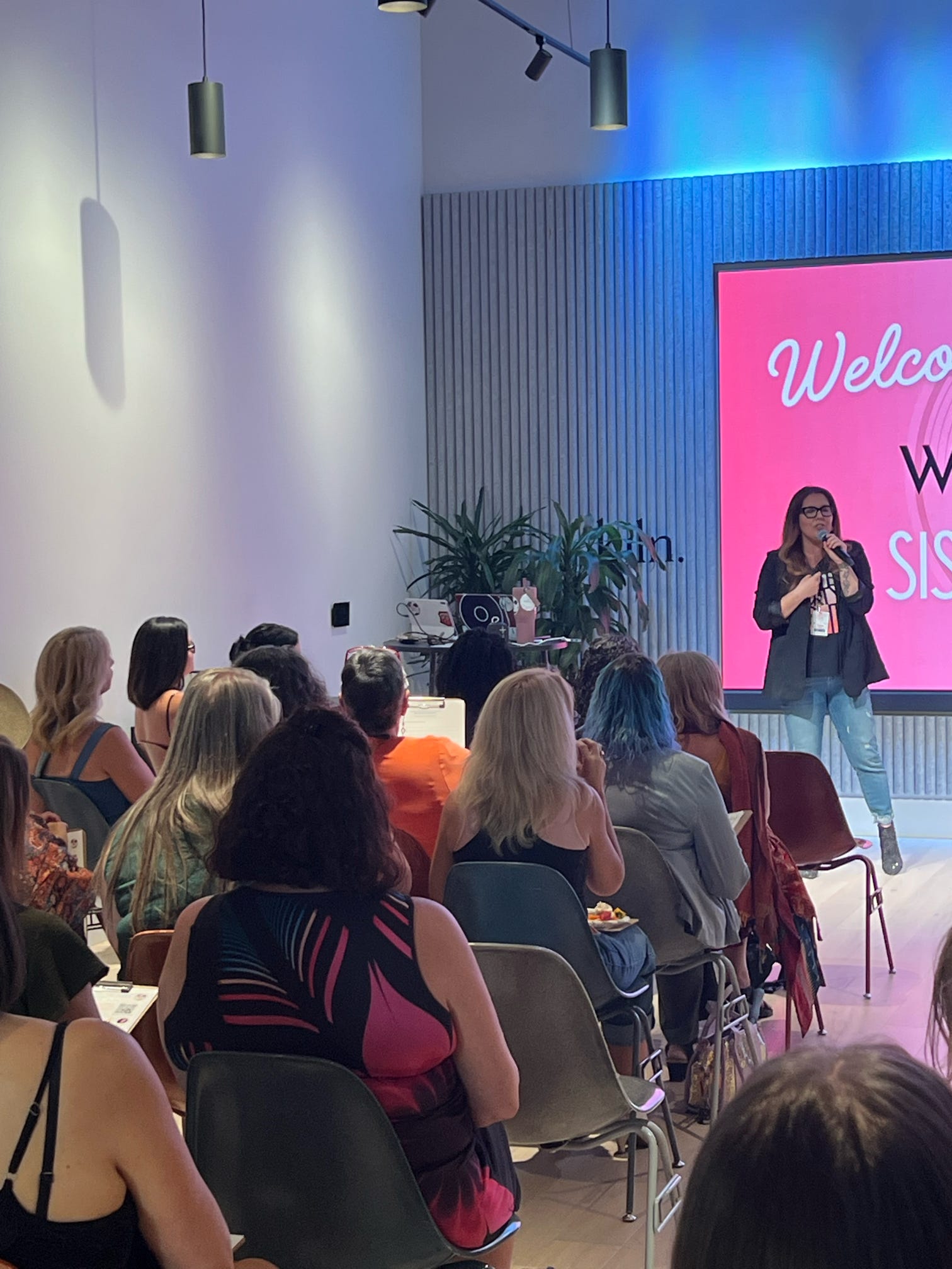A woman speaking on stage in front of an audience in a modern room with a large pink screen that says 'Welcome'.
