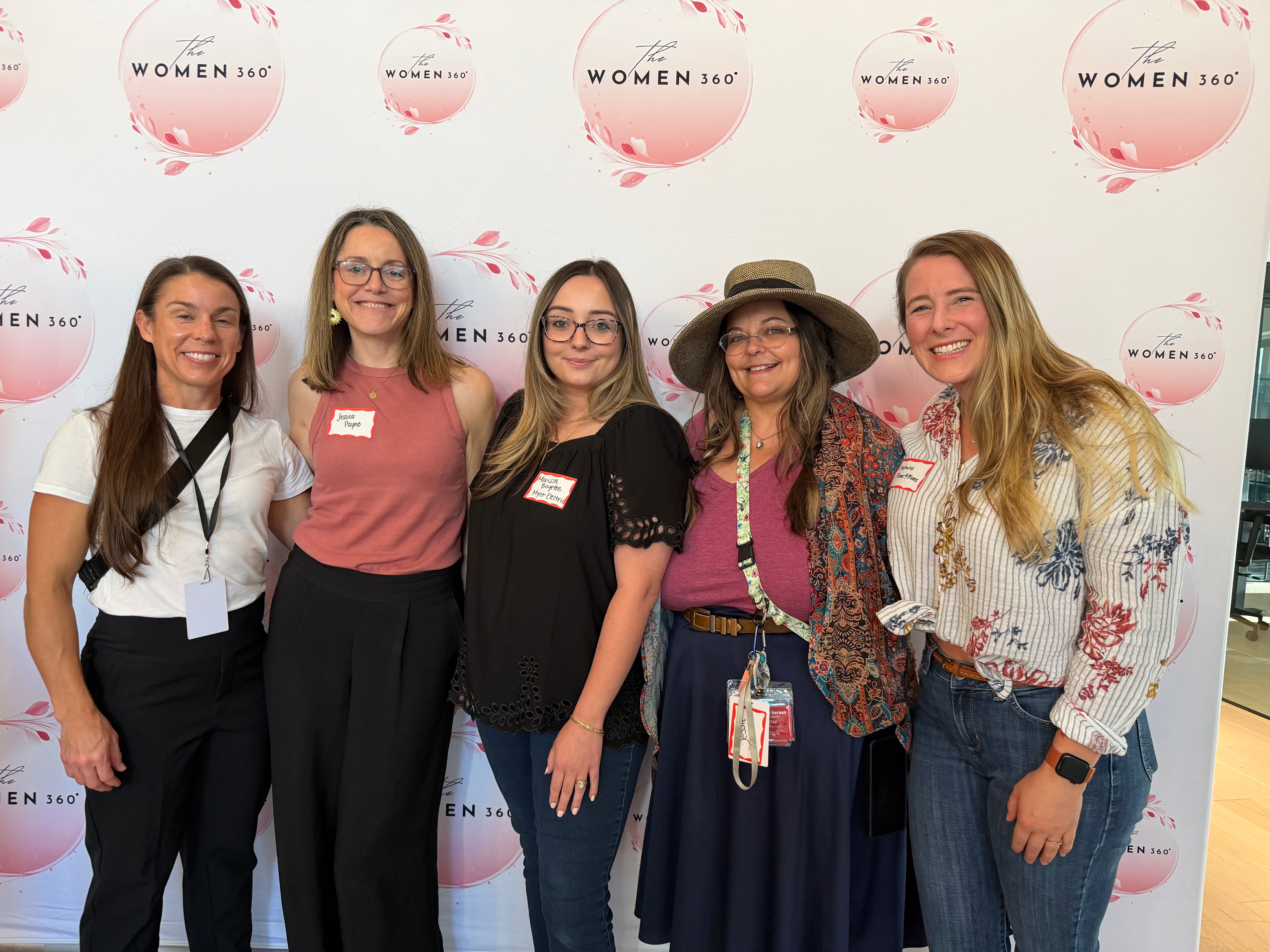 Five women standing together in front of a backdrop with pink circles and text, smiling for the photo.
