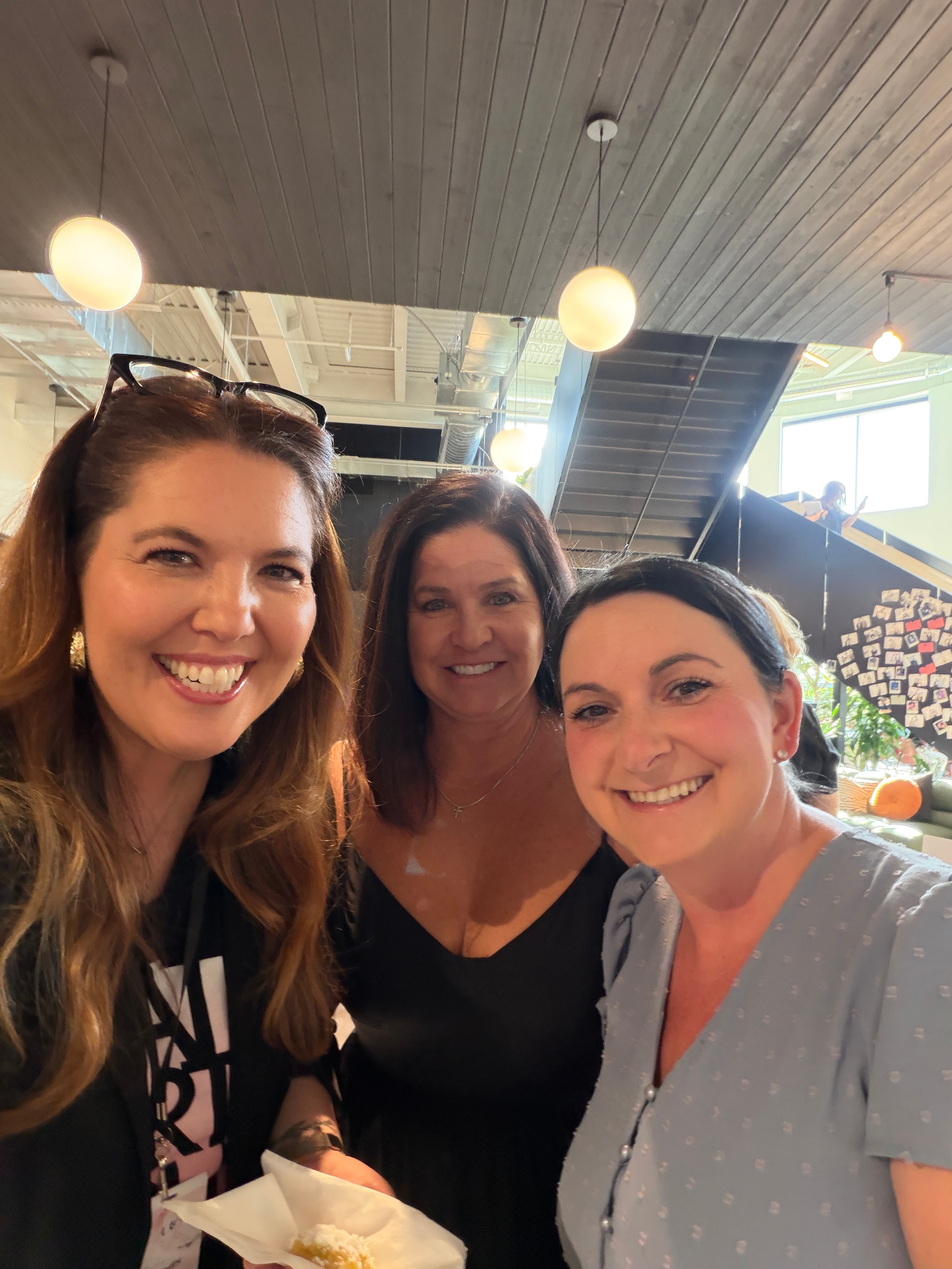 Three women smiling indoors with ceiling lights and windows in the background.