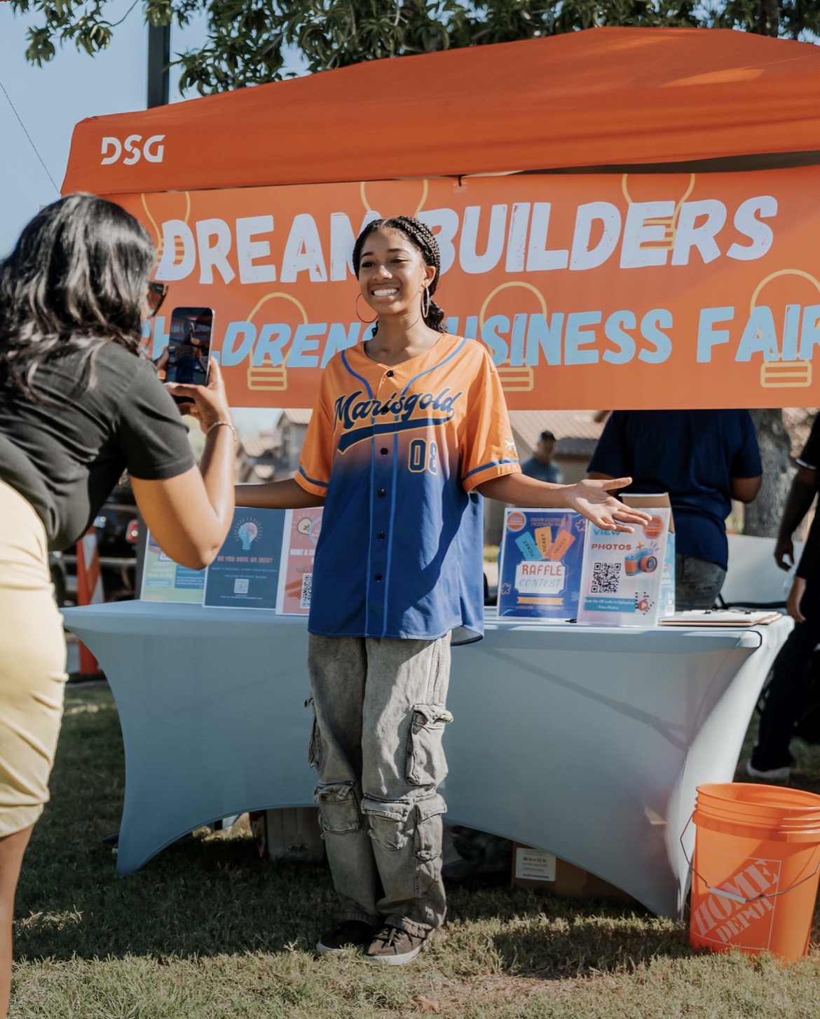 Person smiling and gesturing at an outdoor event with a large orange banner and table, people in the background.