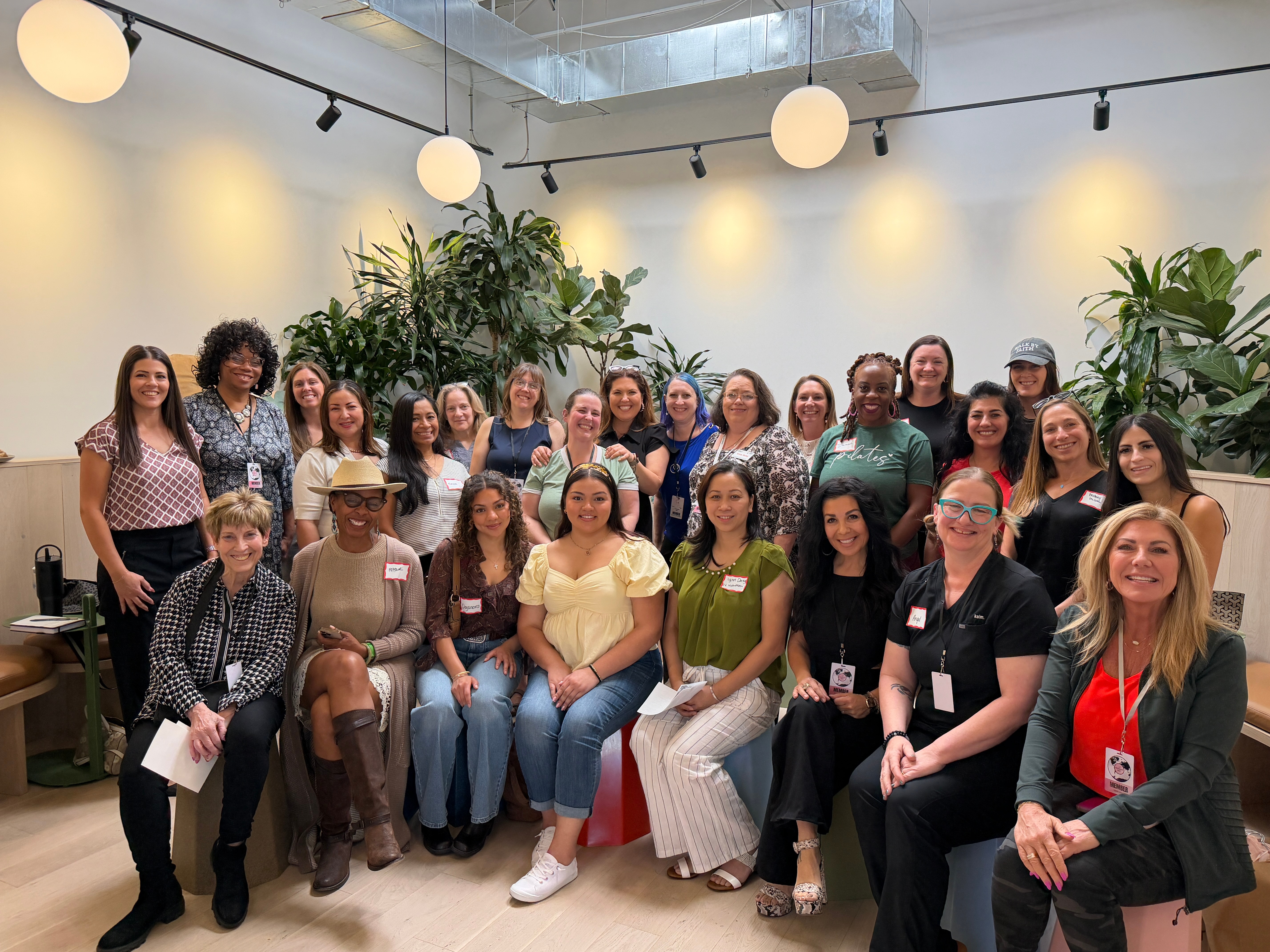 Group of women and one man posing indoors with plants and hanging lights, smiling at the camera.
