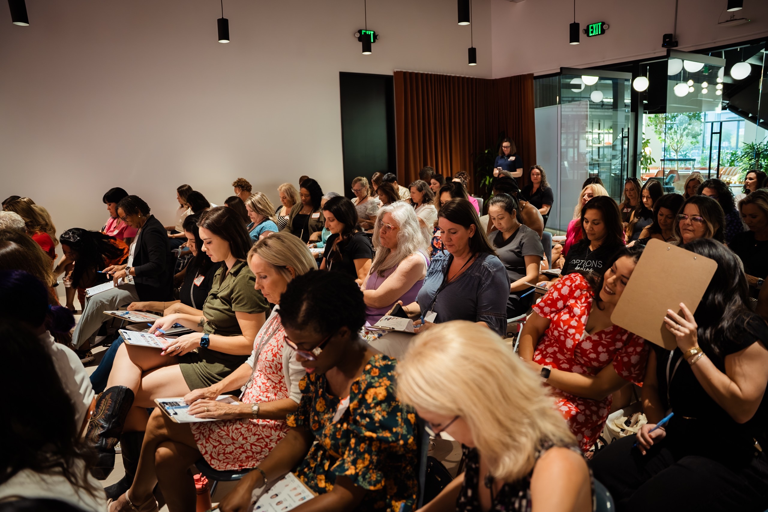 Group of people seated in a room, attending a presentation or event, some taking notes or reading, with a window and curtains in the background.