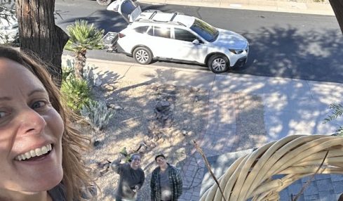 Person smiling outdoors with a view of a street, parked cars, and two people standing on the sidewalk.