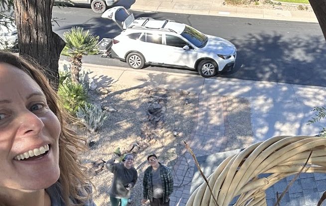 Person smiling outdoors with a view of a street, parked cars, and two people standing on the sidewalk.