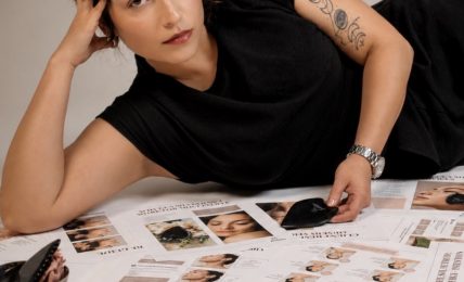 Woman lying on her side on a table covered with magazines, resting her head on her hand, wearing a black top, with tattoos on her arm.