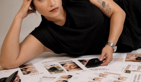 Woman lying on her side on a table covered with magazines, resting her head on her hand, wearing a black top, with tattoos on her arm.