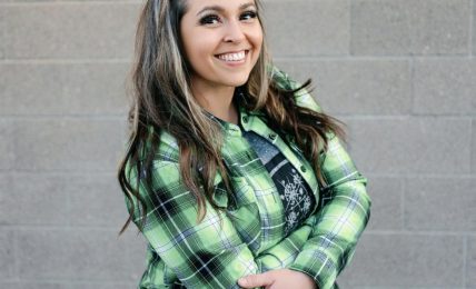 Smiling woman with long hair wearing a green plaid shirt standing against a gray brick wall.